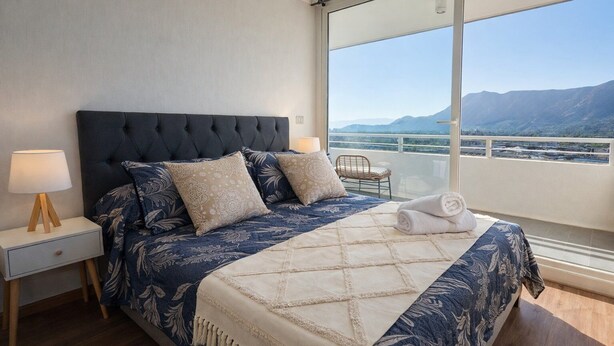 Una camera da letto con un letto grande, un comodino, una lampada e vista sulle montagne attraverso un'ampia finestra.