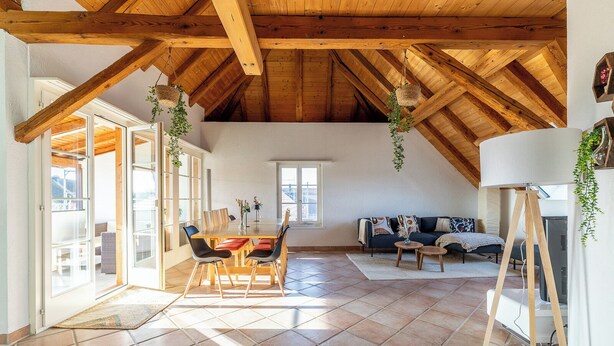 Un loft spacieux avec de hauts plafonds, des poutres en bois, un coin repas avec une table et des chaises, un canapé et une grande fenêtre laissant entrer la lumière naturelle.