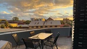 A rooftop terrace with a table and chairs, a covered table, and a view of buildings and a sunset. - 77 on Independence Apartments (Windhoek)