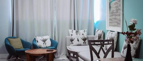Ein Wohnzimmer mit blauem Sessel, rundem Couchtisch, Glastisch mit Vase und einer Couch mit gemusterten Kissen.