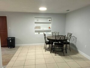 A dining area with a round table and four chairs, a trash can, and a shelf with decorative items. - Entire home in Port Richey, Florida, 2 bedroom 1 bath Residential-Free Parking (Port Richey)