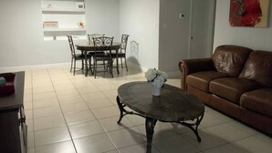 Una sala de estar con un sofá de cuero marrón, una mesa de centro redonda con un jarrón de flores, un comedor con una mesa y sillas, y un estante de pared con objetos decorativos.
