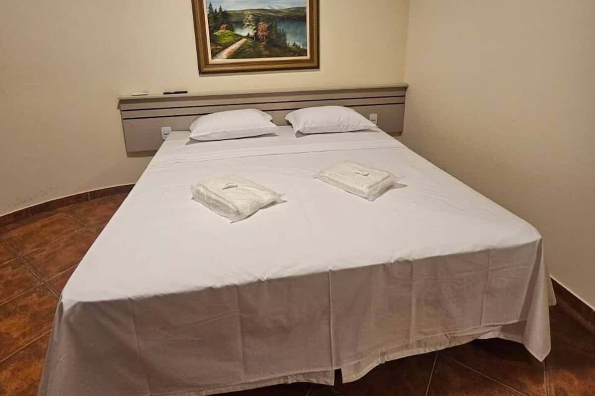 A single-bed room with a white bedspread, two pillows, a nightstand, an air conditioning unit, and a framed landscape painting on the wall.