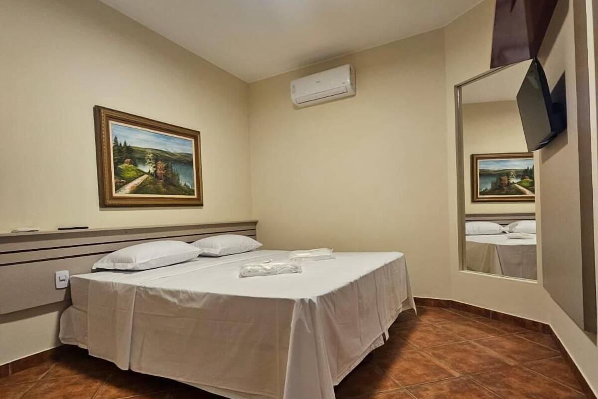 A hotel room with a bed, a painting, an air conditioning unit, and a TV mounted on the wall.