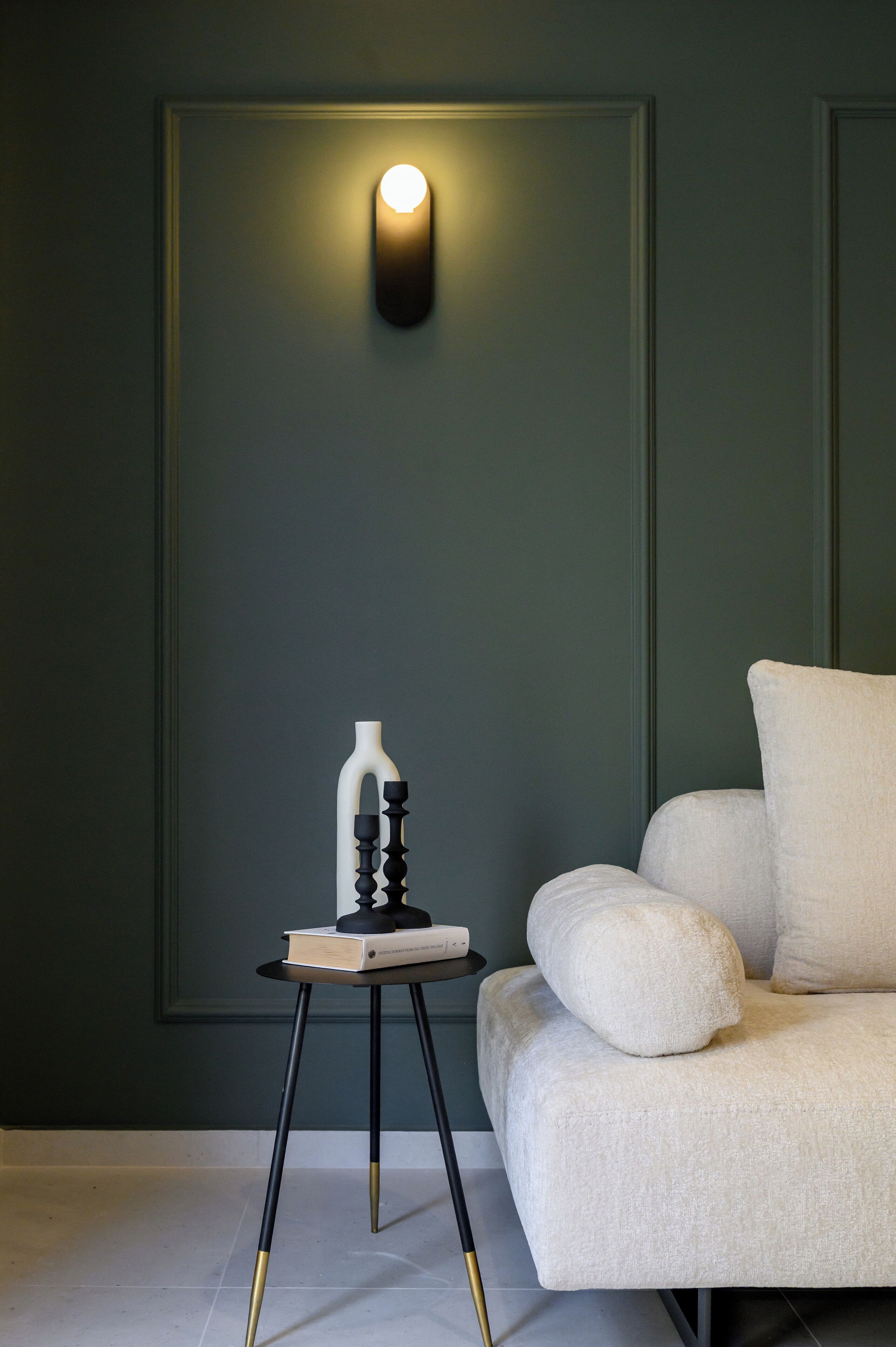 Una habitación moderna con una pared verde oscuro, un sofá blanco con varios cojines, una mesita auxiliar negra y dorada con un libro y un jarrón, y una lámpara de pared.