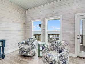 Habitación con paredes de madera, vista al mar, dos sillones con estampado, una mesita y una puerta que da al exterior.