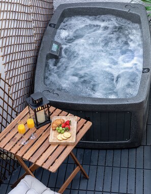 Ein Whirlpool mit Bedienfeld, umgeben von einem Metallzaun, mit einem Holztisch daneben, auf dem Gläser, ein Croissant, Erdbeeren und Weintrauben stehen.