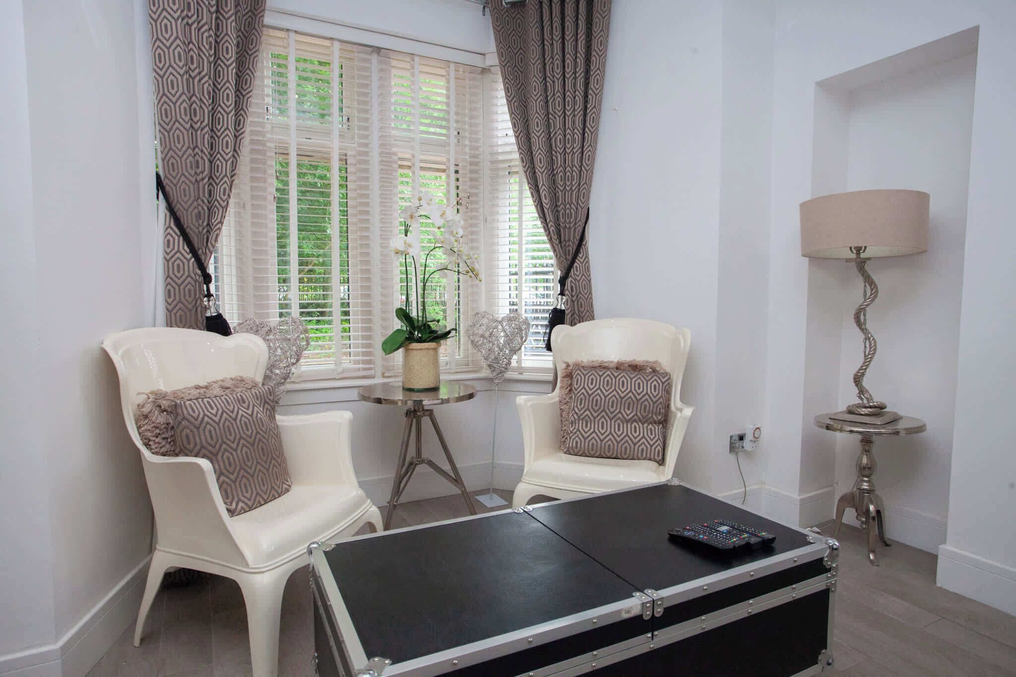 Una habitación con dos sillas blancas, una mesita redonda pequeña, un pouf negro, una lámpara y una ventana con cortinas estampadas.