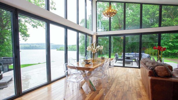 A spacious room with large windows, a wooden floor, a dining table with chairs, and a sofa. - Cameron North Lodge on Loch Lomond (Argyll and Bute Council)