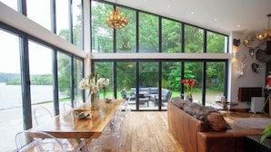 A spacious room with a large wooden dining table, clear plastic chairs, a leather sofa, and a view of a lake through large windows. - Cameron North Lodge on Loch Lomond (Argyll and Bute Council)
