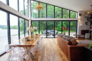 Una habitación amplia con una gran mesa de comedor de madera, sillas de plástico transparente, un sofá de cuero y vistas a un lago a través de amplios ventanales.