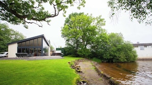 A modern building with a glass facade, a grassy area, and a water body. - Cameron North Lodge on Loch Lomond (Argyll and Bute Council)