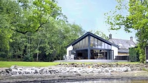 A modern house with a glass facade, a stone retaining wall, and a pond in the foreground. - Cameron North Lodge on Loch Lomond (Argyll and Bute Council)