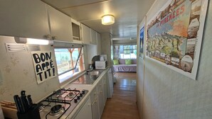 Et lille køkken med gasblus, vask og et vindue med udsigt. Der er et skilt på væggen, der siger "BON APPETIT!", og et billede på væggen, der viser et landskab.