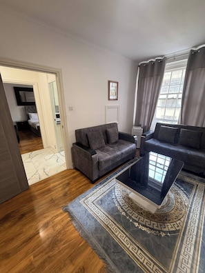 A living room with a grey sofa, a coffee table, and a patterned rug. There is a door leading to a bedroom with a bed and a TV. - Charming Central Apartment Steps From Hyde Park (London)