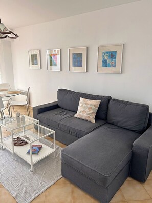 A modern living room with a grey sectional sofa, a glass coffee table, and a white wall with framed artwork. - Les Camelias – Bright One-Bedroom Apartment with Terrace and Garage (Antibes)