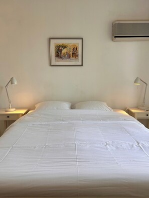 A neatly made bed with white linens, two bedside tables with lamps, a framed picture on the wall, and an air conditioning unit above the bed. - Les Camelias – Bright One-Bedroom Apartment with Terrace and Garage (Antibes)