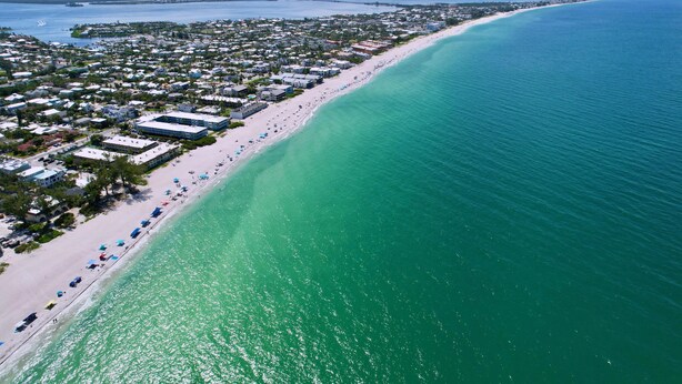 An aerial view of a coastal town with a long sandy beach, clear turquoise waters, and a dense cluster of buildings along the shore. - Sweet Slice of Old Florida Charm with modern amenities, large yard & beach gear (Holmes Beach)