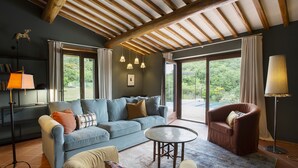 A modern living room with a blue sofa, a brown armchair, and a round coffee table. There are large windows offering a view of greenery outside. - Casa Pernice - Stylish Chianti Retreat with Pool & Vineyard Views (Gaiole in Chianti)