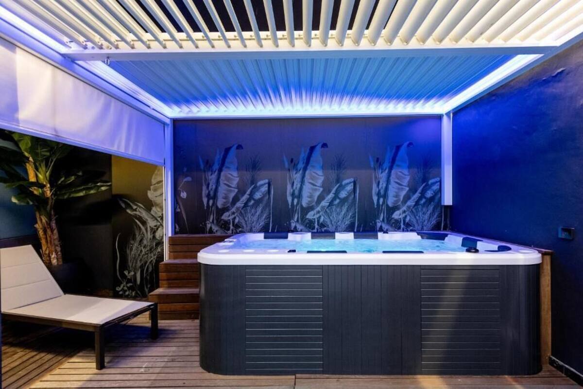 A hot tub with a wooden deck and steps leading up to it, surrounded by a blue-lit wall and a ceiling with white slats.