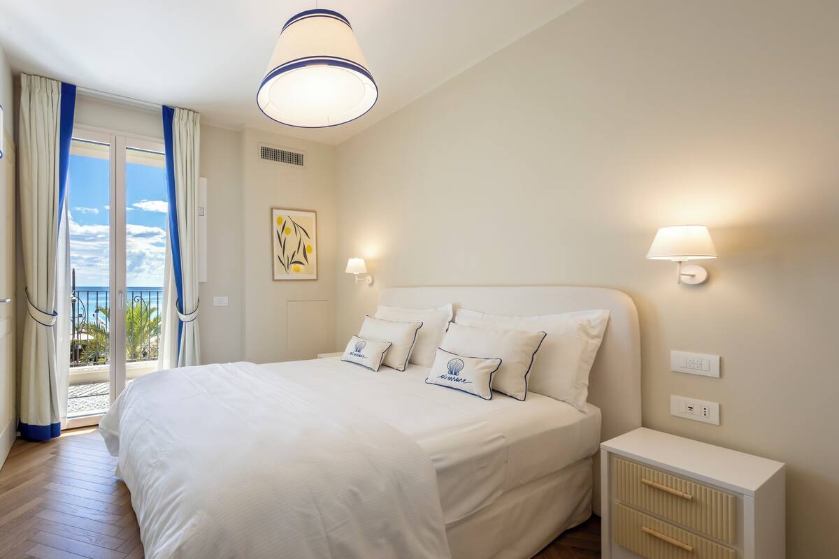 A bedroom with a large bed, two bedside lamps, a nightstand, and a view of the sea through a sliding door.