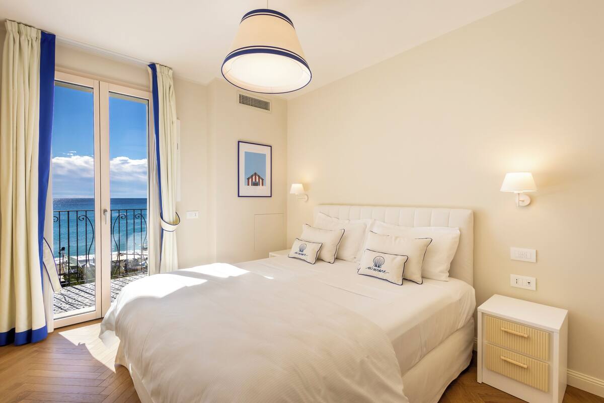 A bedroom with a large bed, a nightstand, a lamp, a framed picture, and a view of the sea through a sliding door.