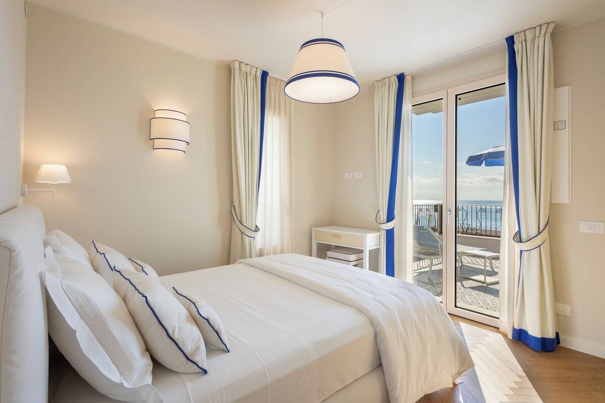 A bedroom with a bed, a desk, and a view of the beach through a sliding door.