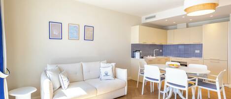 A modern living room with a white sofa, a dining table with chairs, and a kitchen area in the background.