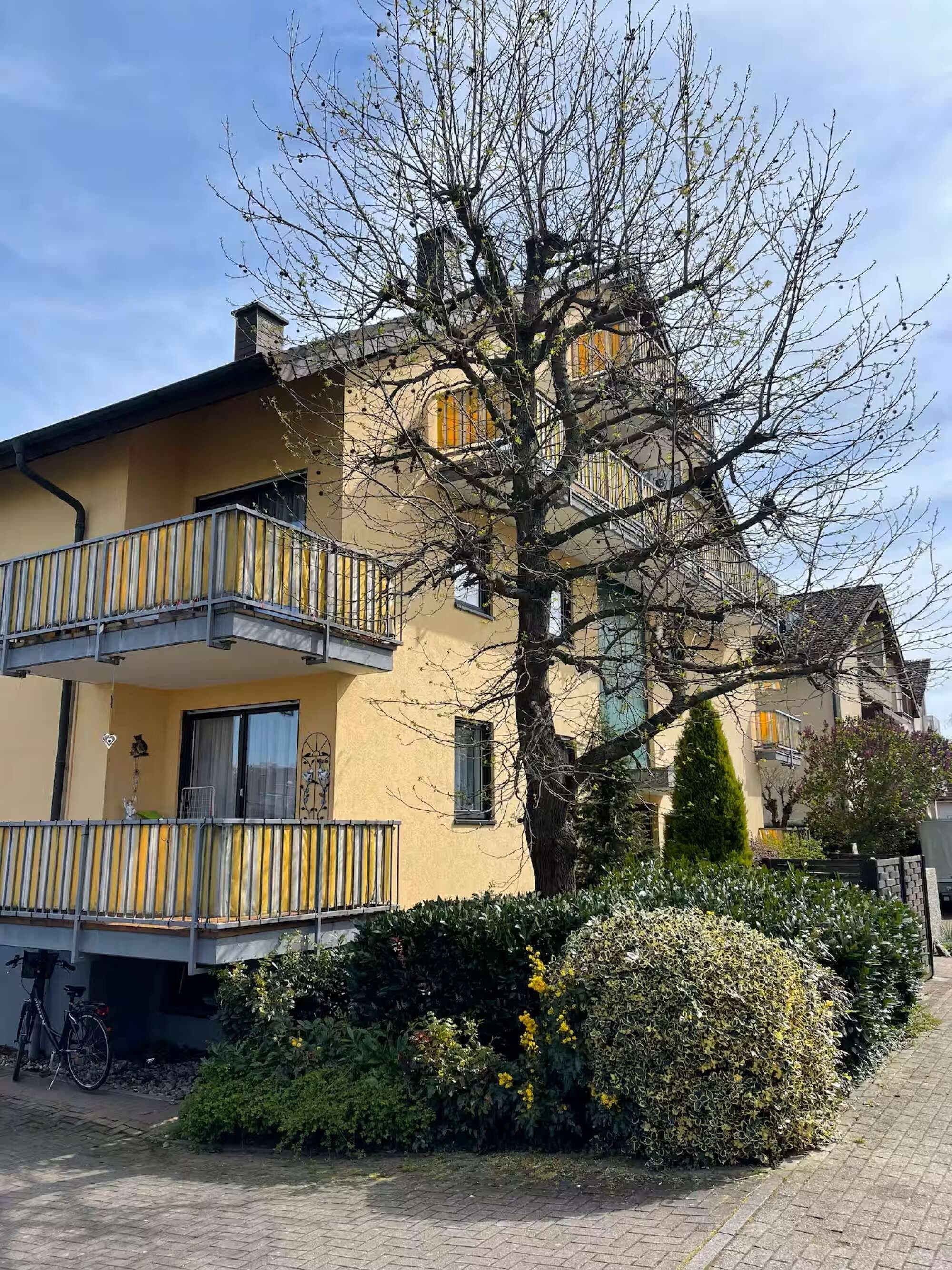 Un immeuble de deux étages avec un balcon, un arbre devant et un vélo garé à l’extérieur.