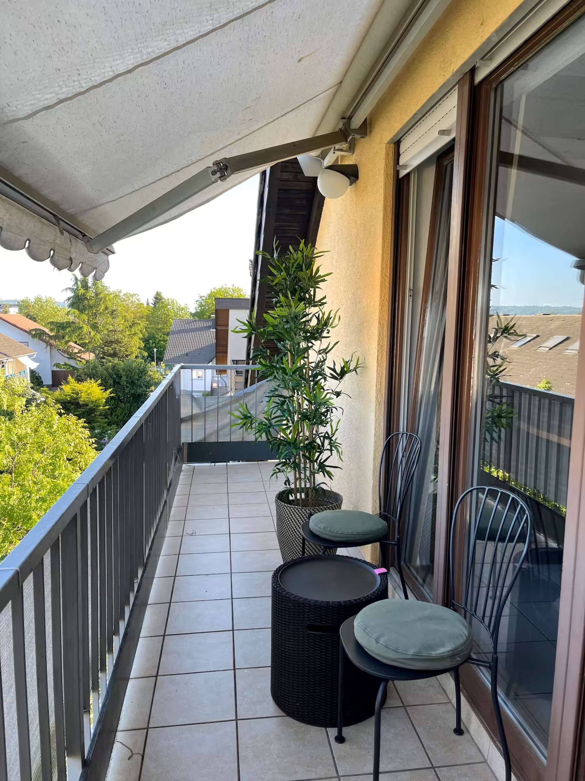 A balcony with a tiled floor, two chairs, a potted plant, and a window with a view of the neighborhood.