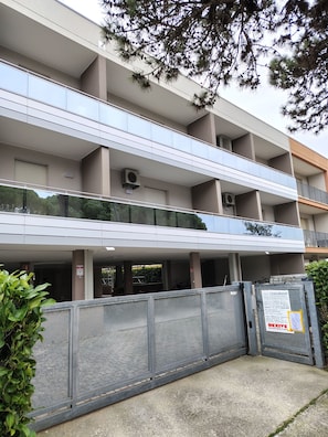 Un immeuble à plusieurs étages doté d’un parking couvert, de climatiseurs et d’une entrée sécurisée par un portail.