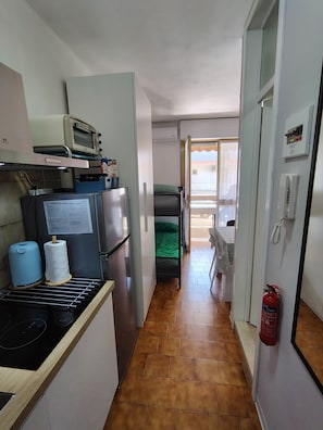 Een compacte keuken met een broodrooster, een koelkast en een kleine oven. Een smalle gang leidt naar een slaapkamer met een groene dekbedovertrek.