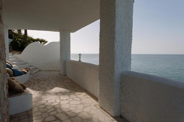 A covered outdoor seating area with a view of the sea, stone flooring, and white walls.