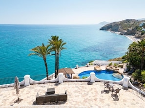 Zona de piscina con vistas al mar, palmeras y un paisaje montañoso.