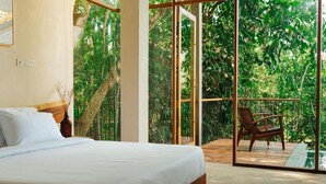 Ein Schlafzimmer mit einem großen Bett, einem Holzstuhl und einer Glastür, die auf eine Terrasse mit Blick auf dichtes Grün führt.