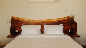 Ein ordentlich bezogenes Bett mit Holz-Kopfteil, zwei Nachttische mit Flaschen und eine schlichte Wand mit Lichtschalter.