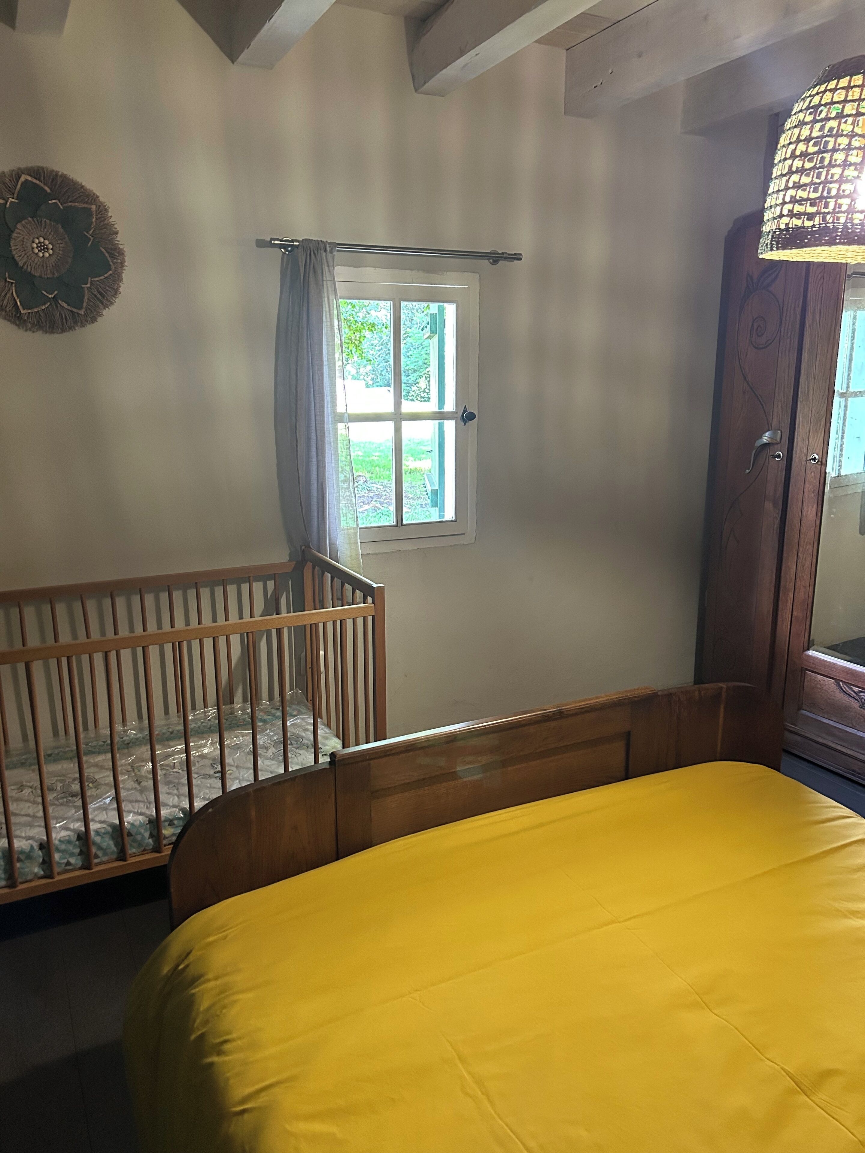 Une pièce avec un berceau en bois, un lit avec un matelas jaune, une fenêtre donnant sur la verdure et une armoire en bois.