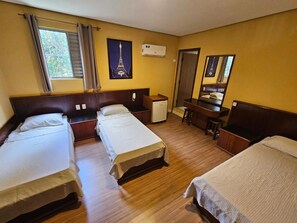 Habitación de hotel con tres camas individuales, un escritorio con silla, un refrigerador pequeño y una ventana con cortinas.