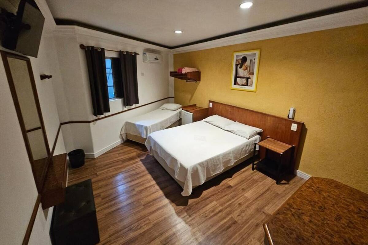 A hotel room with two beds, a wooden floor, a window with curtains, a wall-mounted shelf, and a painting.