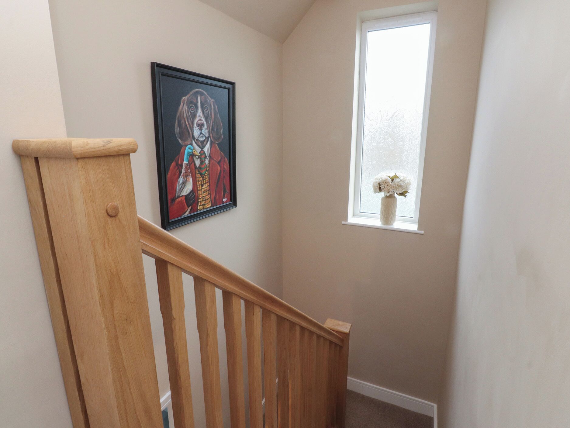 A staircase with a wooden handrail, a framed picture on the wall, and a window with a white vase.