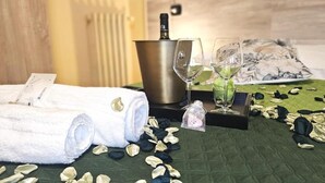 A neatly arranged room with a bed, a bottle of champagne, two wine glasses, a towel, and scattered rose petals.