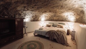 Un dormitorio con techo que recuerda a una cueva, una cama con pétalos de rosa, un armario de madera y una pequeña mesa con lámpara.