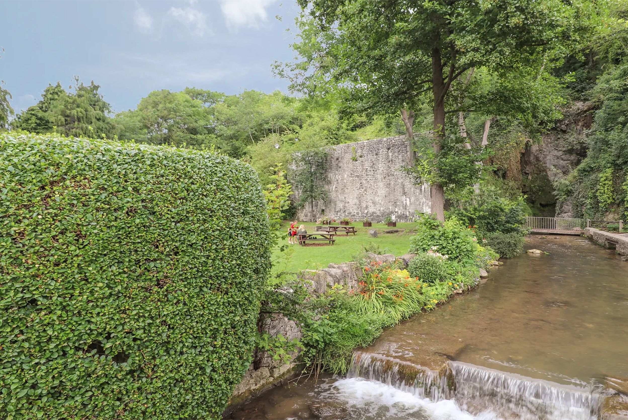 A lush garden with a small waterfall, a stone wall, and a picnic area.