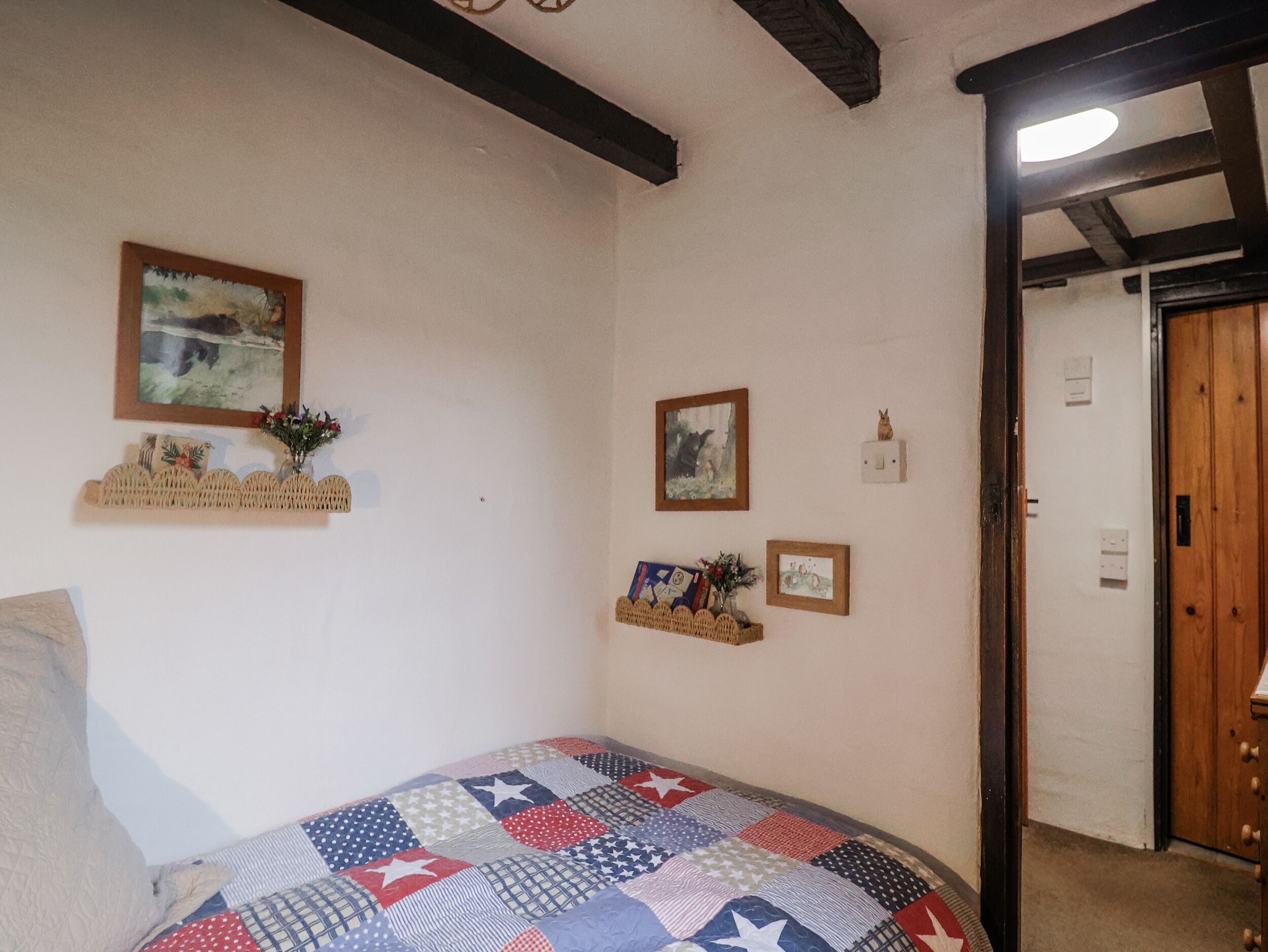 A bedroom with a bed, a quilt, a wooden door, and wall decorations including framed pictures and a shelf with flowers.