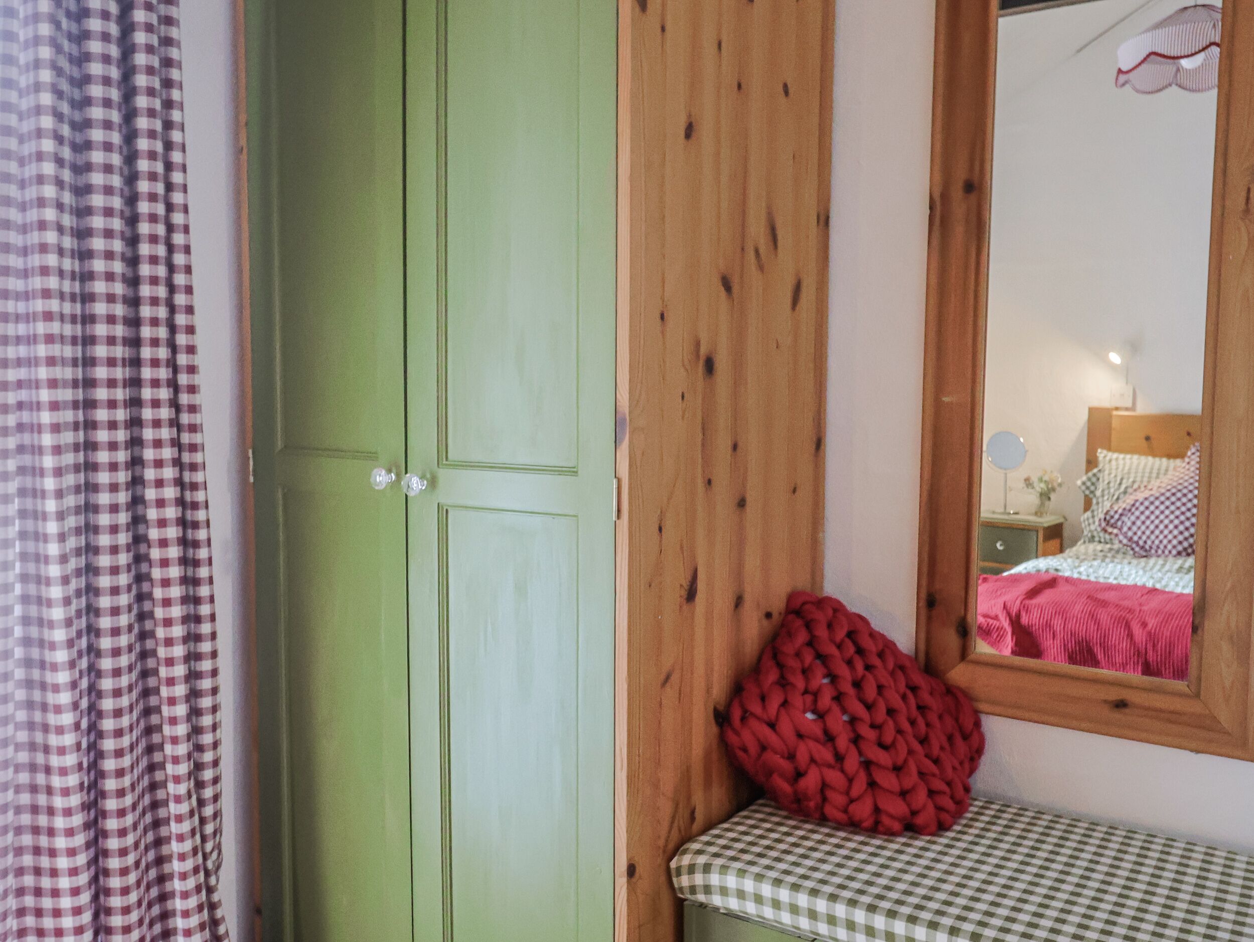 Una stanza con un'armadiatura verde, una panchina a scacchi e uno specchio che riflette un letto con una coperta rosa.