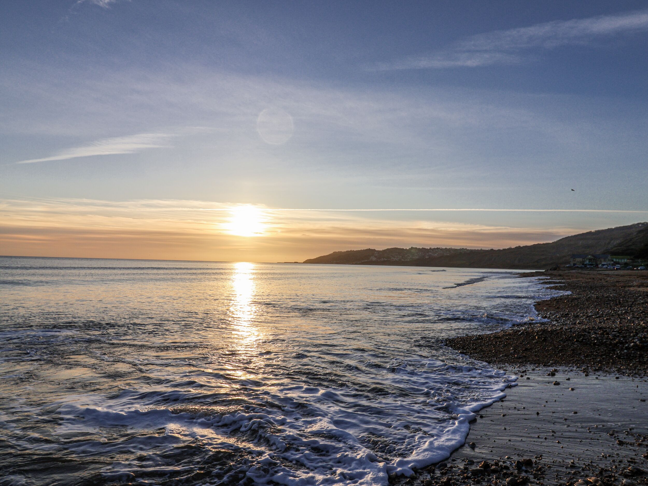 Une plage où l’on peut admirer le coucher de soleil sur l’océan, une côte rocheuse et une colline lointaine.