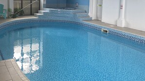 Ein Innenpool mit blauen Mosaikfliesen, einer gefliesten Liegefläche und einer Treppe, die auf eine höhere Ebene führt.
