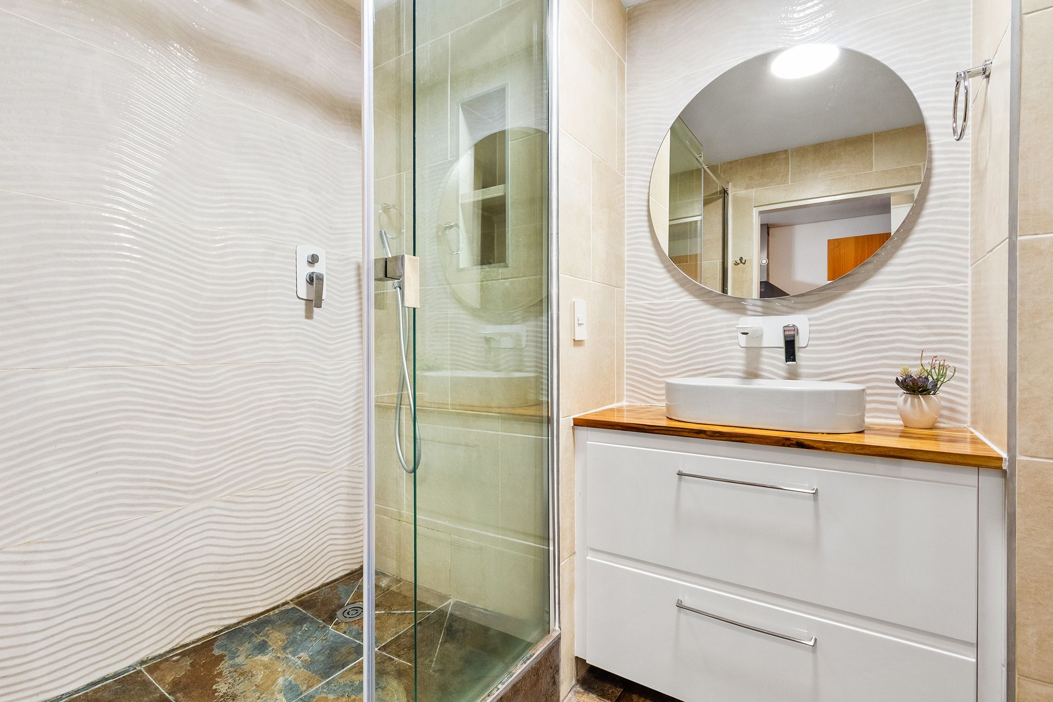 A bathroom with a glass shower enclosure, a round mirror, a sink with a wooden countertop, and a white cabinet.