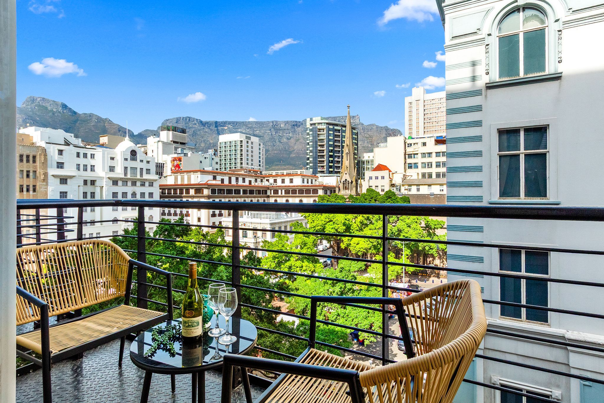A balcony with wicker chairs and a table, overlooking a cityscape with buildings and a mountain in the background.