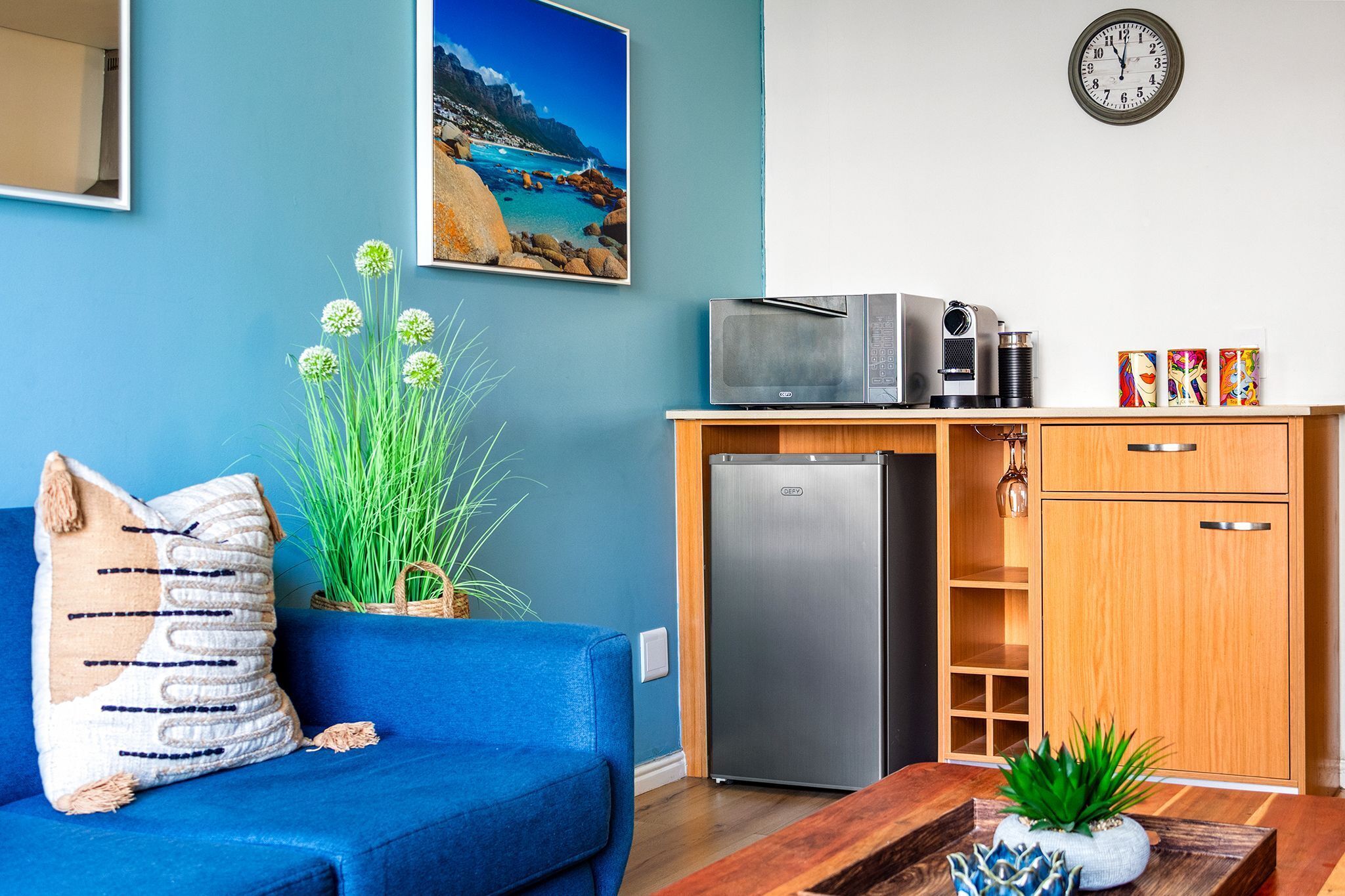 Un salon avec un canapé bleu, un petit coin cuisine comprenant un réfrigérateur, un four à micro-ondes et une horloge murale.